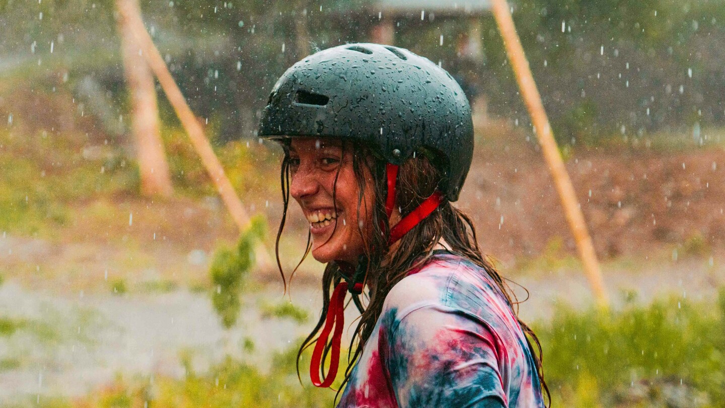 Anna Pixner, eine Downhill-Skateboarderin, steht lächelnd im Regen und trägt einen schwarzen Helm sowie ein buntes, gebatiktes T-Shirt. Im Hintergrund ist eine ländliche Umgebung mit einem Pavillon und mehreren Personen zu sehen.