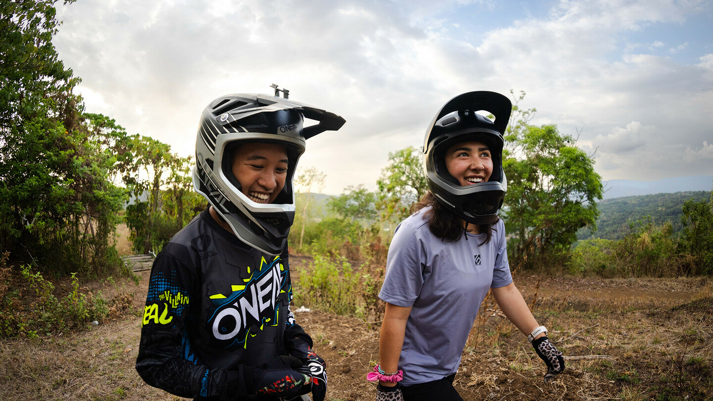 Two young women wearing full-face helmets are smiling in an outdoor setting with trees in the background. The person on the left is wearing a dark jersey, and the person on the right is wearing a purple shirt and spotted gloves.