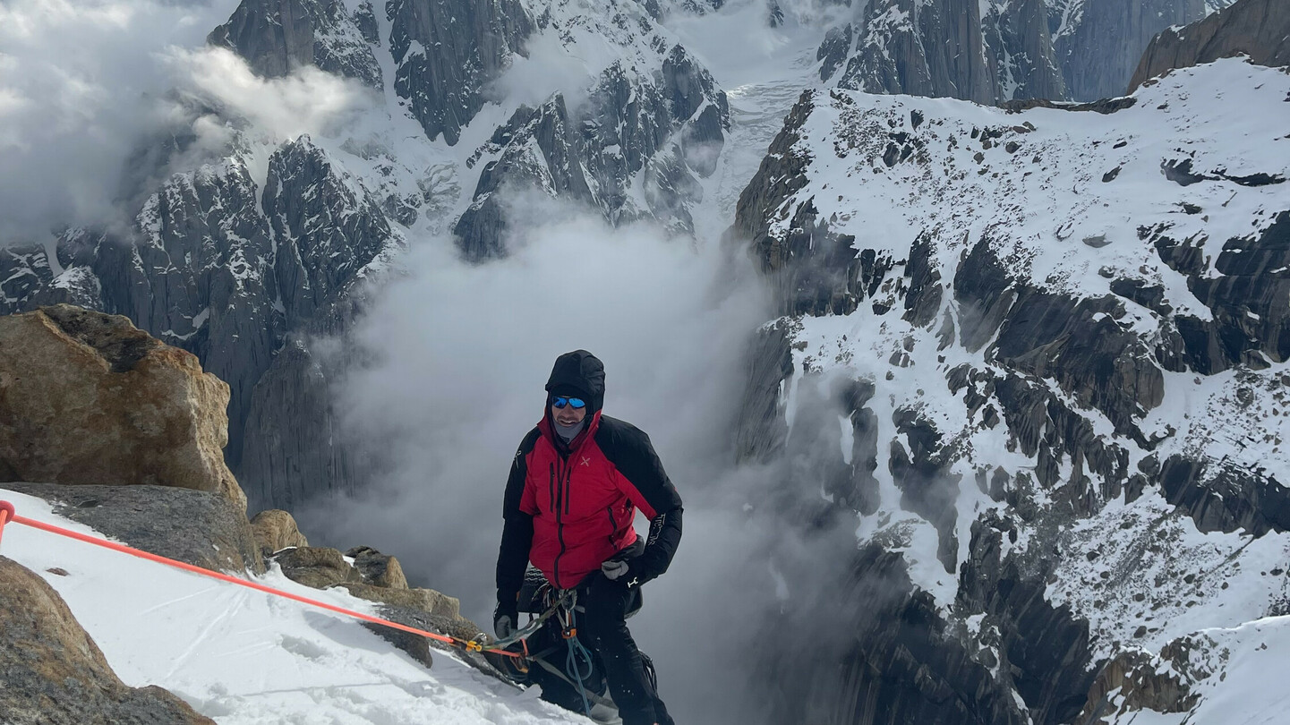 Bergsteiger in roter Jacke beim Aufstieg auf einem verschneiten Hang, umgeben von dramatischen Wolken und schroffen Berggipfeln.