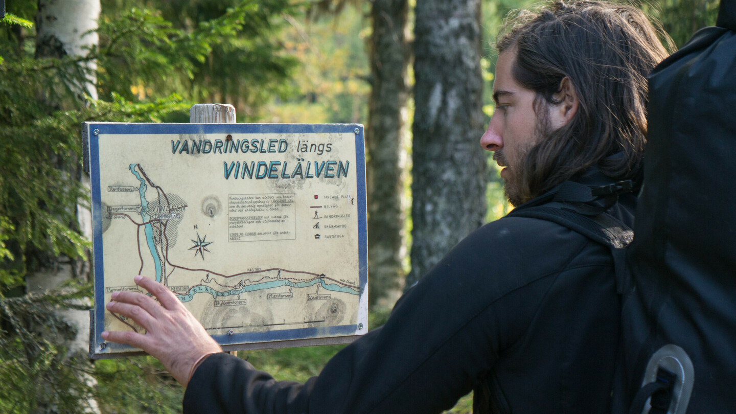 Ein Mann mit schulterlangem Haar und einem grossen Rucksack studiert eine Wanderkarte mit der Aufschrift „Vandringsled längs Vindelälven“ in einem Wald. Er berührt die Karte mit der Hand und wirkt konzentriert. Um ihn herum sind Bäume und dichtes Waldgebiet.