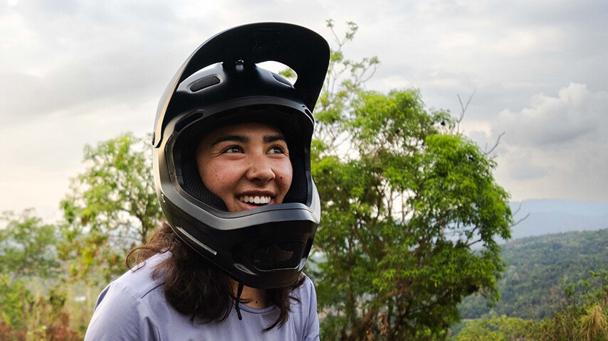 Samantha Soriano l&auml;chelnd mit einem schwarzen Fullface-Helm im Freien. Im Hintergrund sind B&auml;ume und ein bewaldetes H&uuml;gelgebiet zu sehen, der Himmel ist leicht bew&ouml;lkt.
