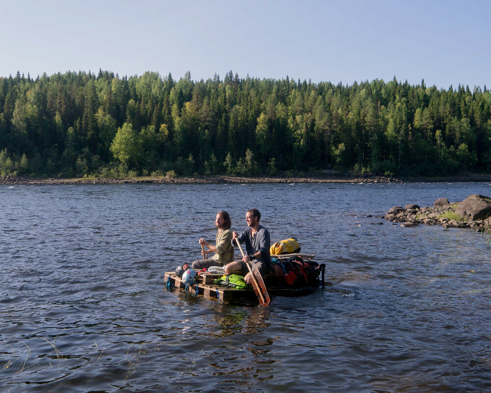 Two people sitting on a makeshift raft made of wooden pallets, drifting on a river surrounded by forested banks. They are holding paddles, with a dense forest visible in the background.